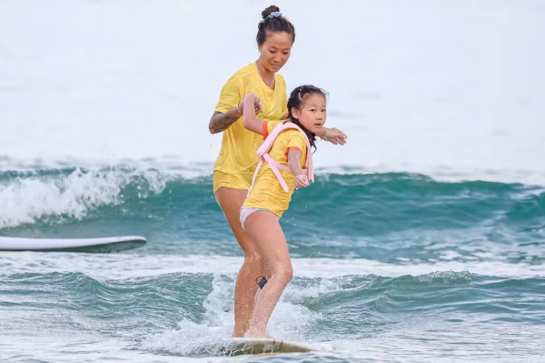 青少年儿童在冲浪教练的陪同下，在三亚市海棠区夏日水上运动嘉年华湾“乘风破浪”。三亚市体育旅游协会供图